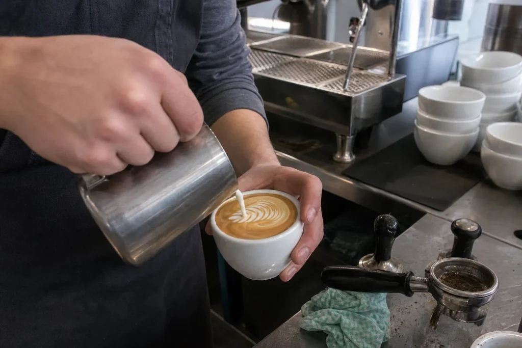 Barista häller upprört mjölk i en espressokopp under en AeroPress VM tävling, med enkel utrustning i bakgrunden.