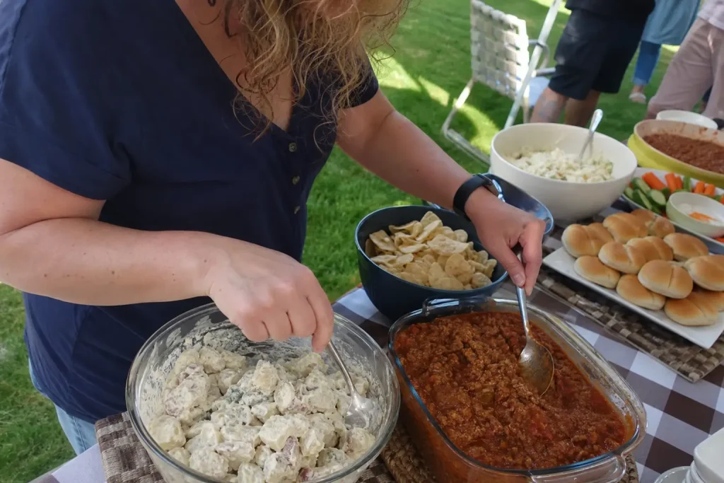 Person tar potatissallad och köttsås med skilda skedar vid buffébord, relevant för så länge midsommarmaten kan stå framme ...