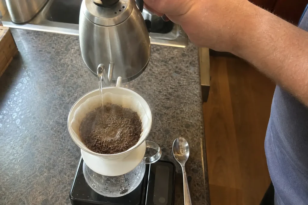 Hand häller hett vatten över kaffesump i dropparbryggare, ett exempel på kaffesmaker och trender i hemmabryggning.