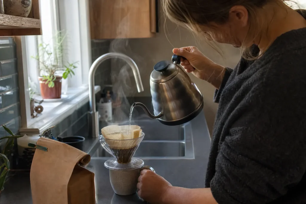 Händer häller varmt vatten i en pour over-tratt vid köksbänken, med kaffebönor och AeroPress VM i närheten.